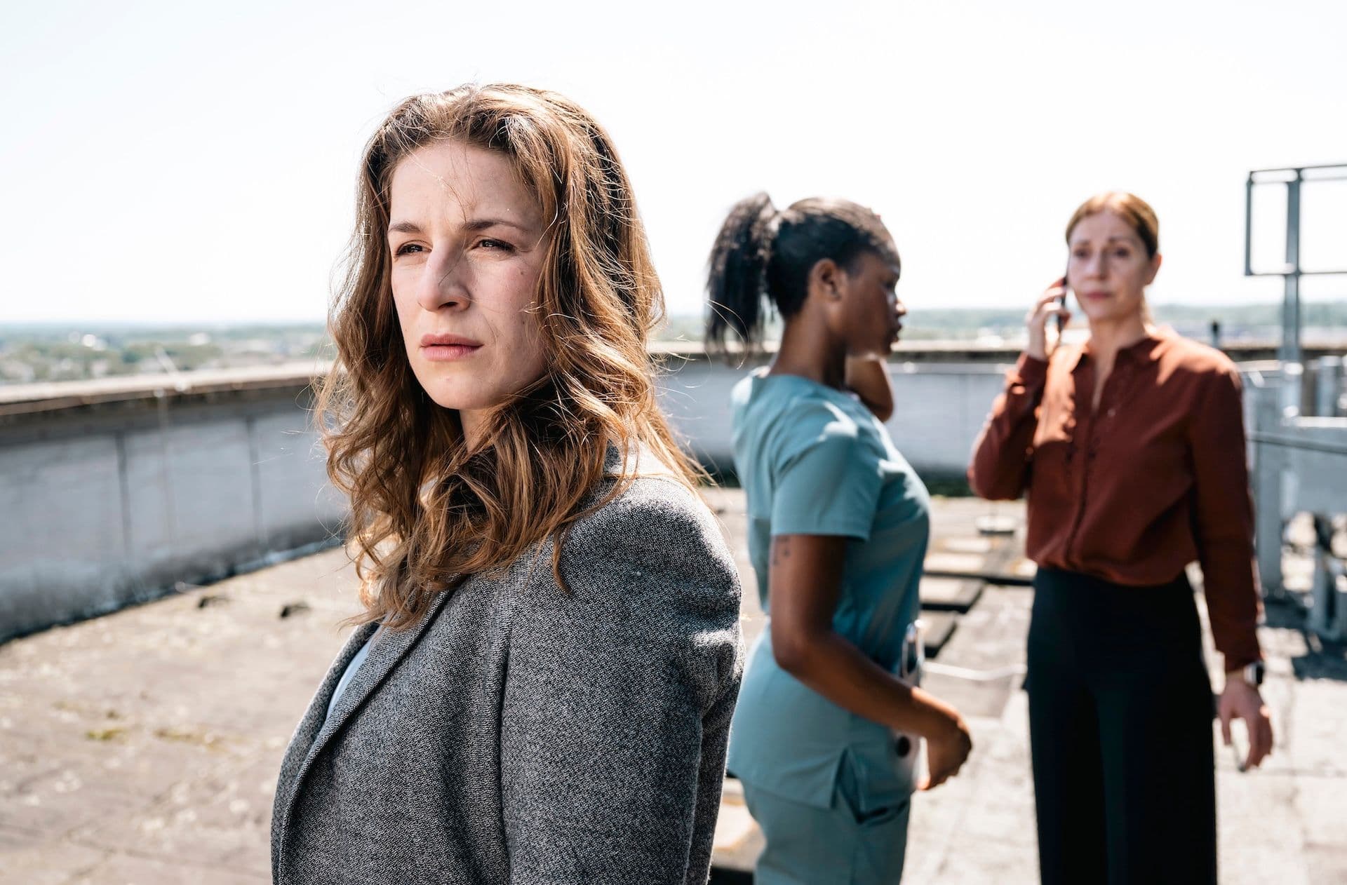 A woman in a gray blazer looks off to the side with a serious expression. | Bildquelle: Three women stand on a rooftop overlooking a city skyline, their expressions suggesting a moment of contemplation or concern. A woman in a gray blazer looks off to the side with a serious expression.
